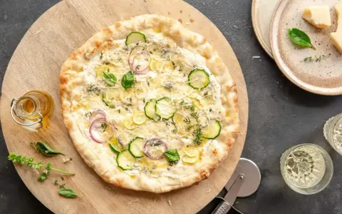 Pyszna pizza bianca z cukinią, cebulą i ziołami, podana na drewnianej desce. Obok buteleczka oliwy i kieliszki z winem.