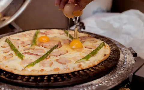Ktoś dodaje jajko do pizzy z boczkiem i szparagami. Pyszna pizza z jajkiem w przygotowaniu.