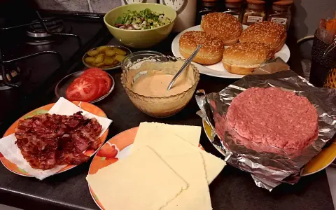 Składniki do przygotowania idealnego cheeseburgera: bułki, surowe mięso, ser, bekon, pomidory, ogórki i sos.