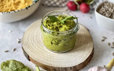 Pesto z liści rzodkiewki w słoiczku, ozdobione natką pietruszki, na drewnianym krążku. Obok czosnek, rzodkiewki i nasiona.