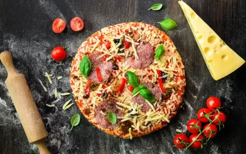 Gotowa pizza z salami, serem i warzywami, obok wałek, pomidorki i ser. Idealna na wieczór z przyjaciółmi, gdy brakuje czasu na przygotowanie ciasta z mąki na pizzę.