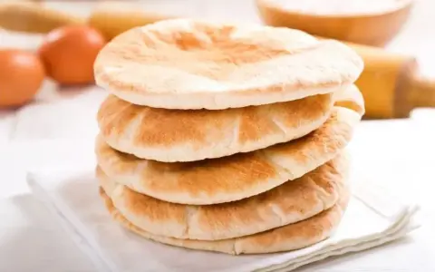 Stos świeżo upieczonego chleba arabskiego na białej serwetce, obok jajka i wałka do ciasta.