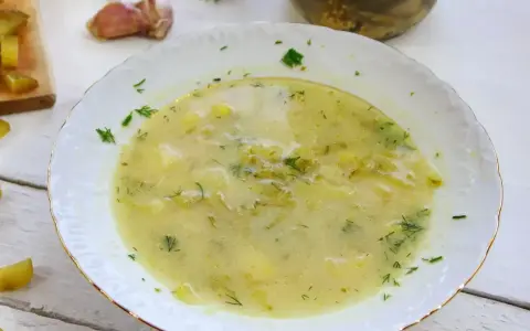 Aksamitna zupa ogórkowa z ziemniakami i koperkiem, podana w białej misce ze złotym rantem. Obok czosnek i ogórki kiszone.