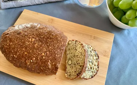 Chleb z twarogu, bogaty w ziarna i orzechy, pokrojony na desce. Obok miska z winogronami i szklanka wody.
