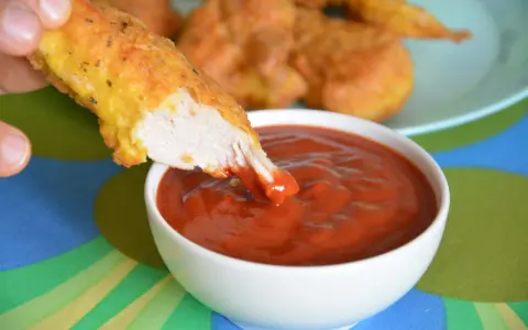 Chrupiący nugget zanurzany w gęstym, czerwonym sosie do nuggetsów. Na talerzu w tle widać więcej złotych kawałków kurczaka.