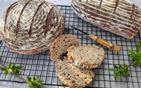 Dwa bochenki chleba żytniego na zakwasie, jeden okrągły, drugi podłużny, z kilkoma kromkami na ruszcie.