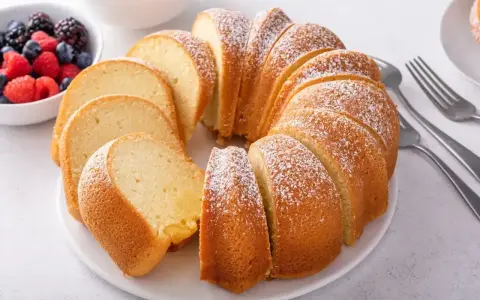 Babka piaskowa przepis babci, puszysta, pokrojona, posypana cukrem pudrem, obok miseczka z owocami i widelce.