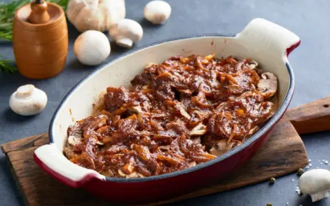 Karkówka pieczona w plastrach z cebulą i sosem, przypominającym ketchup. Danie w naczyniu żaroodpornym, obok grzyby i czosnek.