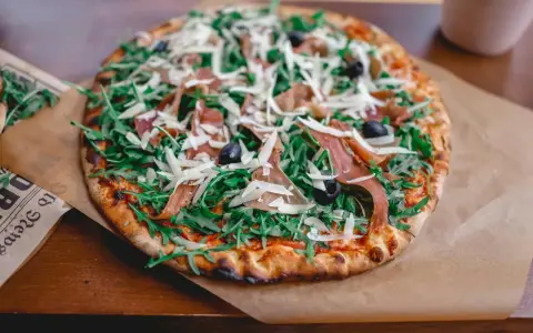 Apetyczna pizza prosciutto z rukolą, oliwkami i parmezanem, podana na papierze.