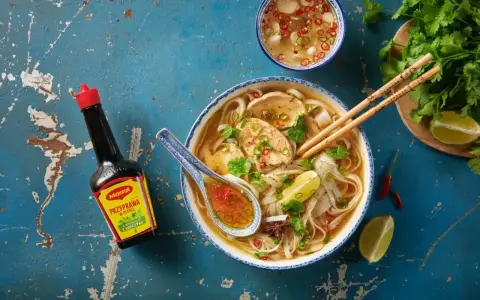 Aromatyczny makaron pho z kurczakiem, świeżą kolendrą, limonką i papryczkami chili. Obok butelka sosu Maggi.