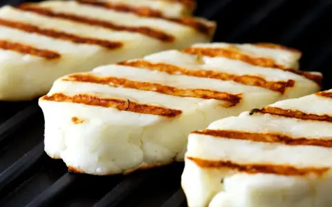 Trzy kawałki halloumi z grilla, z charakterystycznymi brązowymi paskami, leżą na czarnej, grillowej powierzchni.