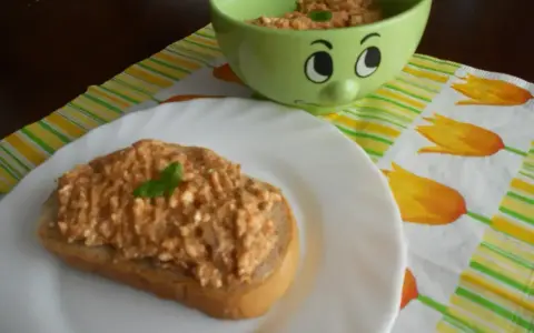 Pyszna pasta z makreli i twarogu na kromce chleba, udekorowana listkiem bazylii. Obok miseczka z pastą i wesołą buźką.