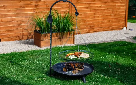 Grill trójnóg z paleniskiem i rusztem, na którym znajdują się kiełbaski, pieczywo i warzywa.
