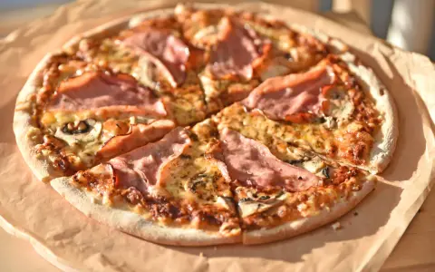 Pizza z szynką i pieczarkami, idealna na szybki obiad. Jakie pieczarki do pizzy? Te ciemne, pokrojone w plasterki, świetnie komponują się z serem i wędliną.