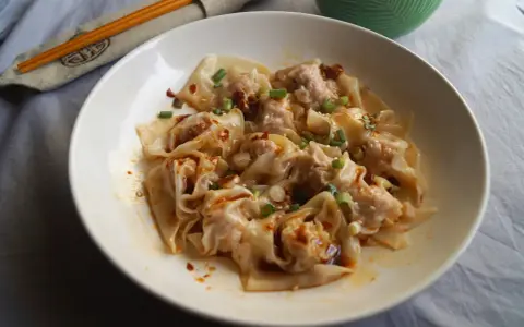 Pyszne ciasto wonton polane pikantnym sosem, posypane szczypiorkiem i sezamem. Obok pałeczki i miseczka z sosem.
