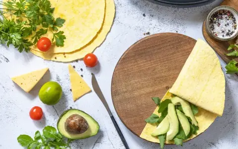 Keto tortilla z awokado i rukolą na drewnianej desce, obok pomidorki, ser i limonka.