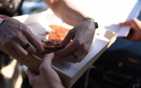 Dwie osoby dzielą się pizzą z pudełka. Właśnie pokazują sobie, jak jeść pizzę, by każdy dostał swój kawałek.