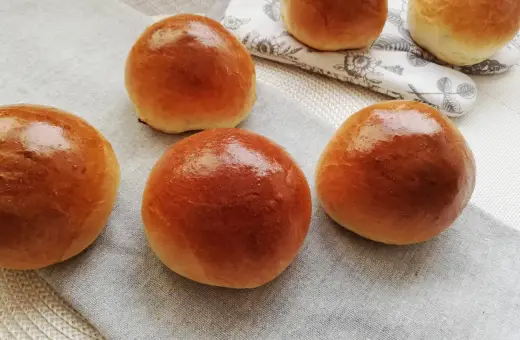 Pięć puszystych bułek maślanych, lśniących od pieczenia, ułożonych na lnianej serwetce.