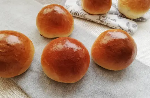Pięć puszystych bułek maślanych, lśniących od pieczenia, ułożonych na lnianej serwetce.