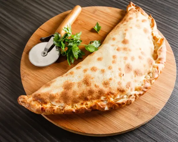 Złocisty calzone, czyli włoska pierożkowa pizza, leży na desce z krążkiem do pizzy i natką pietruszki.
