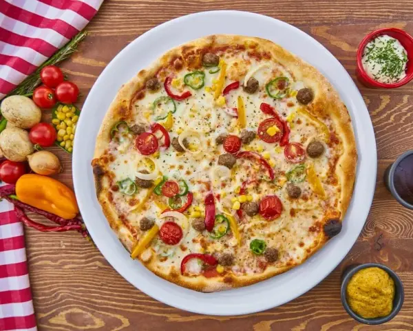 Pizza z mięsem mielonym, pomidorkami, papryką i kukurydzą na białym talerzu, obok sosy i warzywa.