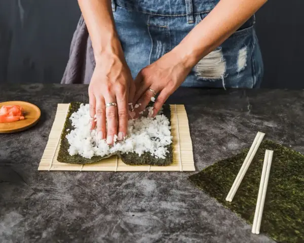Ręce rozkładają ryż na nori, przygotowując zaprawę do sushi na 500g ryżu. Obok leżą pałeczki i imbir.