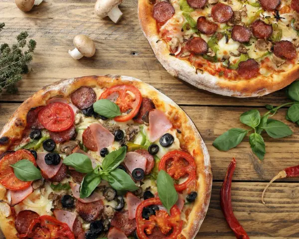 Dwie pizze z dodatkami: salami, pomidory, oliwki, szynka, pieczarki i bazylia.