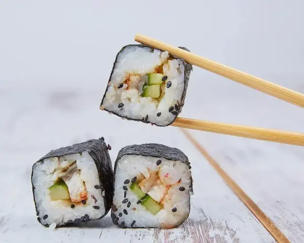 Trzy kawałki sushi z ryżem, ogórkiem i rybą, jeden podniesiony pałeczkami. Idealne do sosu, który stanowi zalewa do sushi.
