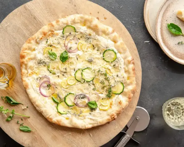 Pyszna pizza bianca z cukinią, cebulą i ziołami, podana na drewnianej desce. Obok buteleczka oliwy i kieliszki z winem.