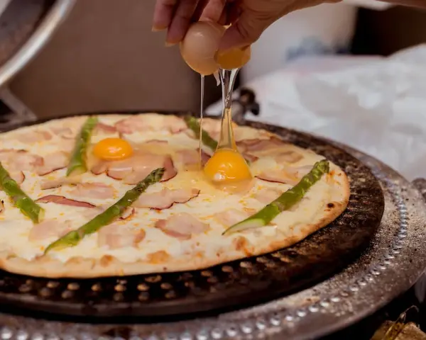 Ktoś dodaje jajko do pizzy z boczkiem i szparagami. Pyszna pizza z jajkiem w przygotowaniu.