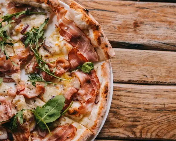 Pyszna pizza carbonara z chrupiącym boczkiem i rukolą, podana na drewnianym blacie.