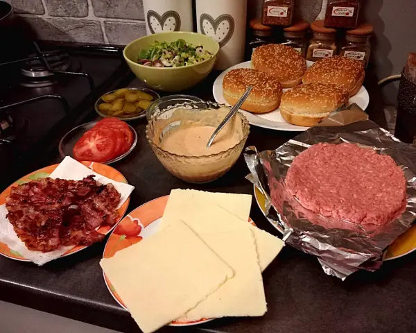 Składniki do przygotowania idealnego cheeseburgera: bułki, surowe mięso, ser, bekon, pomidory, ogórki i sos.