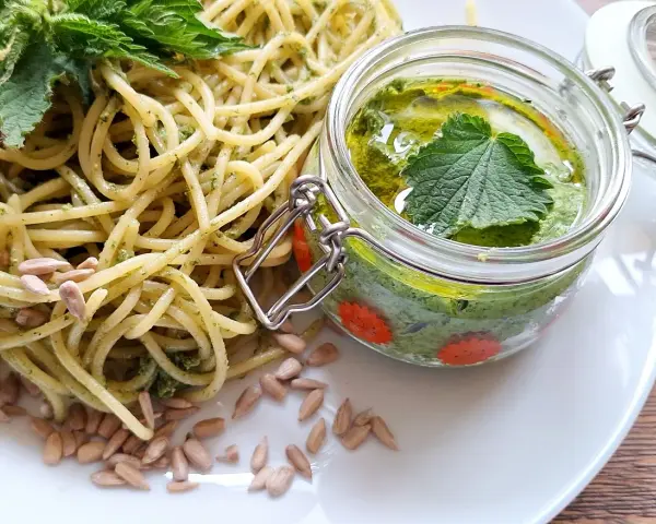 Makaron z pesto z pokrzywy, ozdobiony świeżymi liśćmi pokrzywy i pestkami słonecznika. Obok słoiczek z domowym pesto.