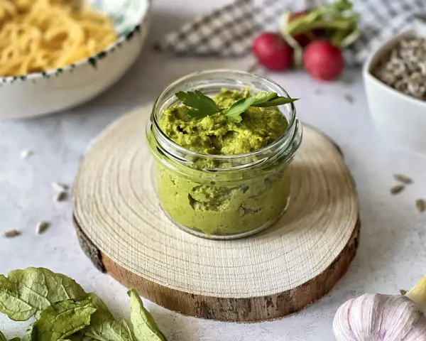 Pesto z liści rzodkiewki w słoiczku, ozdobione natką pietruszki, na drewnianym krążku. Obok czosnek, rzodkiewki i nasiona.