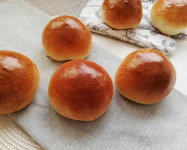 Pięć puszystych bułek maślanych, lśniących od pieczenia, ułożonych na lnianej serwetce.