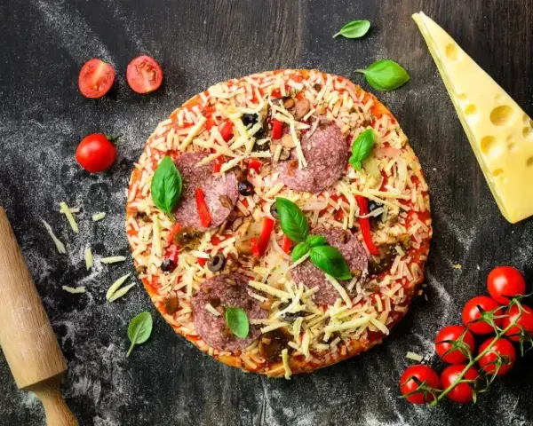 Gotowa pizza z salami, serem i warzywami, obok wałek, pomidorki i ser. Idealna na wieczór z przyjaciółmi, gdy brakuje czasu na przygotowanie ciasta z mąki na pizzę.