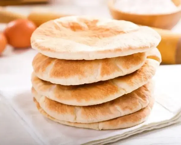 Stos świeżo upieczonego chleba arabskiego na białej serwetce, obok jajka i wałka do ciasta.