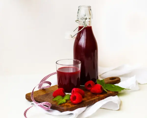 Szklanka i butelka z ciemnoczerwonym sokiem z malin, na drewnianej desce z malinami i listkami mięty.