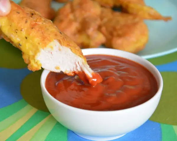 Chrupiący nugget zanurzany w gęstym, czerwonym sosie do nuggetsów. Na talerzu w tle widać więcej złotych kawałków kurczaka.