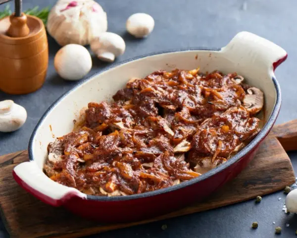 Karkówka pieczona w plastrach z cebulą i sosem, przypominającym ketchup. Danie w naczyniu żaroodpornym, obok grzyby i czosnek.