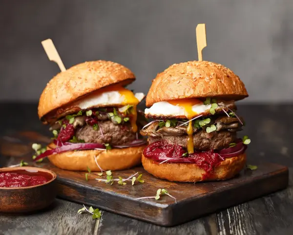 Dwa soczyste burgery z jajkiem sadzonym i grzybami. Idealny stopień wysmażenia burgera podkreśla smak. Obok miseczka sosu.