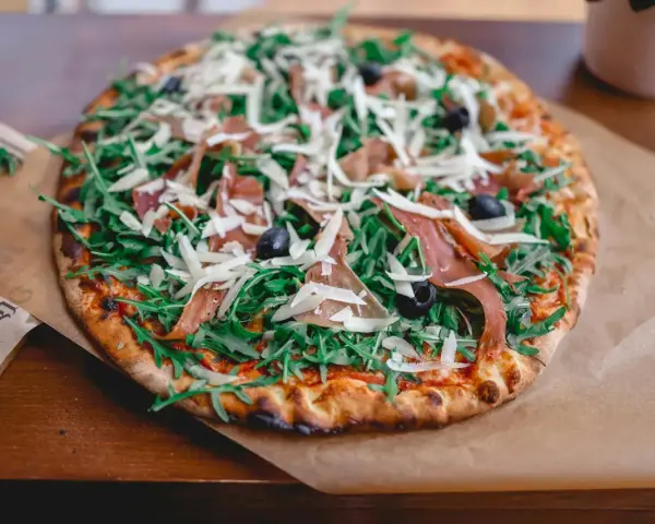 Apetyczna pizza prosciutto z rukolą, oliwkami i parmezanem, podana na papierze.