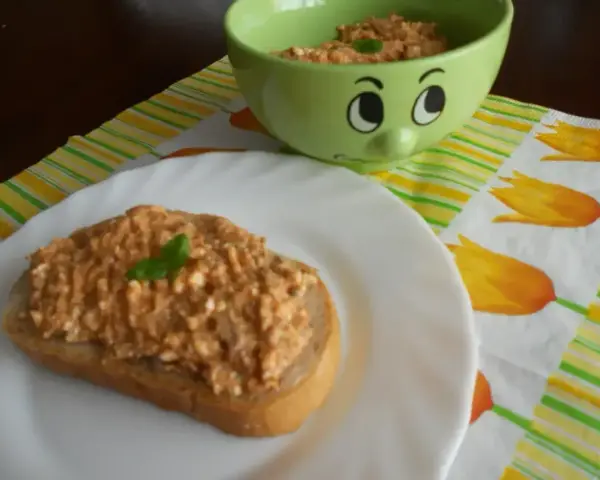 Pyszna pasta z makreli i twarogu na kromce chleba, udekorowana listkiem bazylii. Obok miseczka z pastą i wesołą buźką.