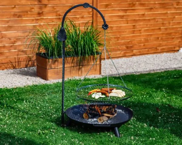 Grill trójnóg z paleniskiem i rusztem, na którym znajdują się kiełbaski, pieczywo i warzywa.