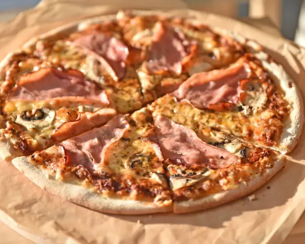 Pizza z szynką i pieczarkami, idealna na szybki obiad. Jakie pieczarki do pizzy? Te ciemne, pokrojone w plasterki, świetnie komponują się z serem i wędliną.