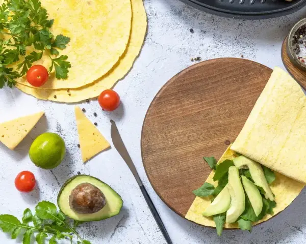 Keto tortilla z awokado i rukolą na drewnianej desce, obok pomidorki, ser i limonka.