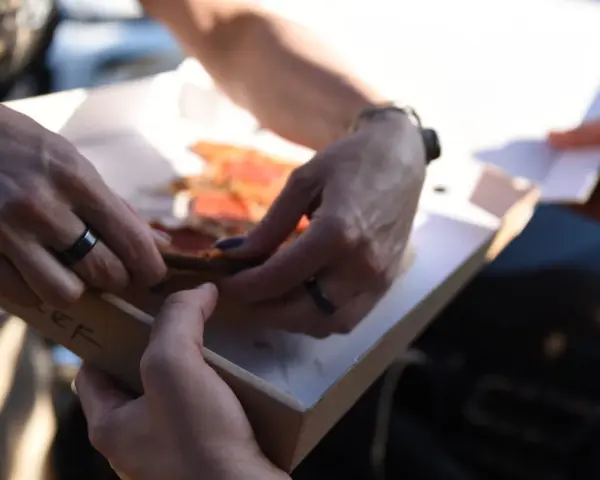 Dwie osoby dzielą się pizzą z pudełka. Właśnie pokazują sobie, jak jeść pizzę, by każdy dostał swój kawałek.