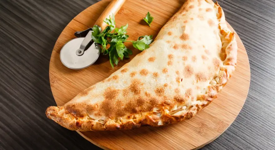 Złocisty calzone, czyli włoska pierożkowa pizza, leży na desce z krążkiem do pizzy i natką pietruszki.