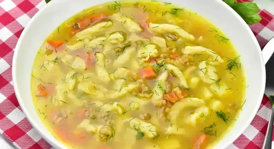 Aromaticzna zupa zacierkowa z kawałkami marchewki i świeżym koperkiem, podana w białej misce na czerwono-białej kratce.