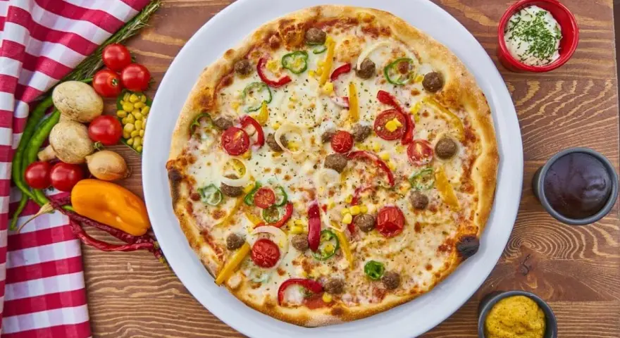 Pizza z mięsem mielonym, pomidorkami, papryką i kukurydzą na białym talerzu, obok sosy i warzywa.